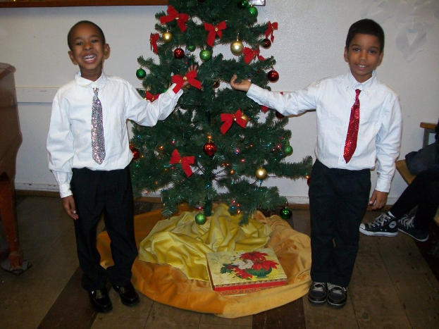 Ein geschmückter Weihnachtsbaum steht in einem Raum mit einem Holztisch links, einem Stuhl rechts und zwei Jungen in weißen Hemden, die lächeln und den Baum halten, mit verschiedenen Gegenständen und einer weißen Wand im Hintergrund.