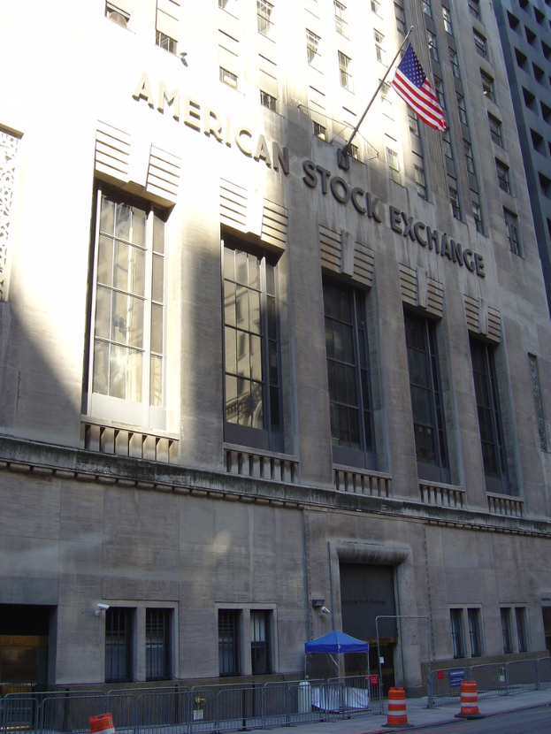 Ein Gebäude mit Text an seiner Fassade, ein Fahnenmast an der Wand, ein Geländer auf dem Gehweg davor, Barrieren jenseits des Geländers und eine Straße im rechten unteren Bereich.