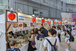 Eine Gruppe von Menschen auf einem Food-Festival in Japan sitzt an einem Tisch mit Essen, einige halten Gläser und tragen Taschen, während andere die Gegenstände auf dem Tisch untersuchen; Tafeln mit Text und Bildern, Lichter und Laternen hängen von der Decke, mit einem Gebäude im Hintergrund.