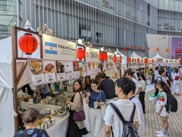 Eine Gruppe von Menschen auf einem Food-Festival in Japan sitzt an einem Tisch mit Essen, einige halten Gläser und tragen Taschen, während andere die Gegenstände auf dem Tisch untersuchen; Tafeln mit Text und Bildern, Lichter und Laternen hängen von der Decke, mit einem Gebäude im Hintergrund.