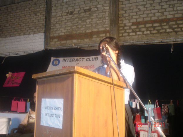 Ein Mädchen in einem weißen Hemd steht neben einem Rednerpult in einem Konferenzraum, mit einem Mikrofonständer rechts daneben, einem weißen Banner auf einem Hintergrund und mehreren Taschen, die an einem Faden rechts hängen.