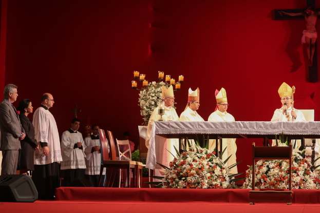 Vier Personen in einem Raum mit Kerzen auf Tischen, eine Person hält ein Mikrofon, Stühle, ein Lautsprecher, Blumen und heilige Kreuze an der Wand im Hintergrund.