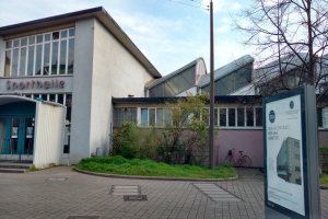 Ein großes Gebäude mit einem Schild davor, umgeben von Bäumen, Pflanzen, Gras, Fahrrädern und einem Pfahl, mit einem bewölkten Himmel im Hintergrund, identifiziert als das Sportballe, das 2020 eröffnet wird.