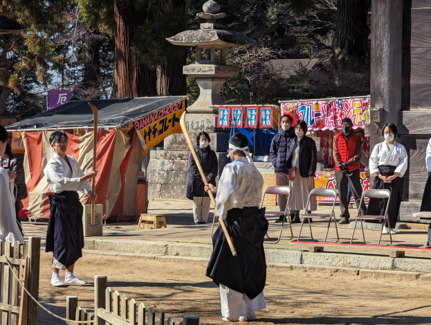 Eine Gruppe von Menschen vor einem Gebäude während einer traditionellen japanischen Zeremonie in Kyoto, einige tragen Masken und halten Stöcke, mit Stühlen, einem Zelt, Bannern, einem Pfahl, einer Säule, Bäumen, einem hölzernen Zaun, einem Seil und einem klaren blauen Himmel im Hintergrund.