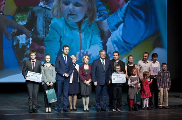 Eine Gruppe von Menschen auf einer Bühne mit Taschen und Urkunden, im Hintergrund ein Bildschirm mit einem Bild eines Kindes und anderen.