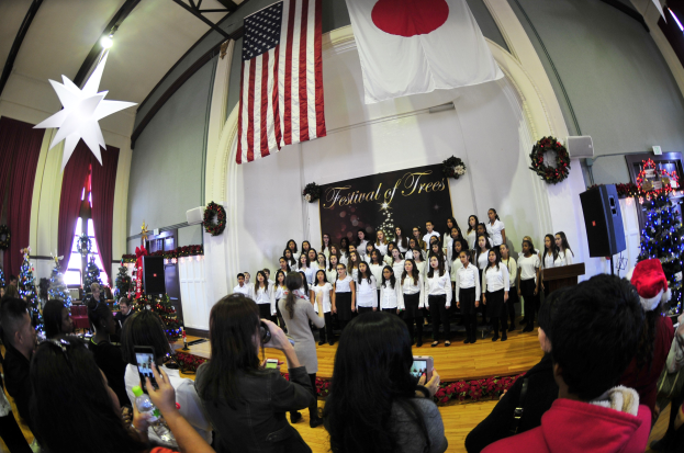 Eine Gruppe von Menschen auf einer Bühne vor einem Publikum mit geschmückten Weihnachtsbäumen, einem Rednerpult, Lautsprechern, einer Texttafel, Vorhängen, Flaggen und Deckenleuchten.