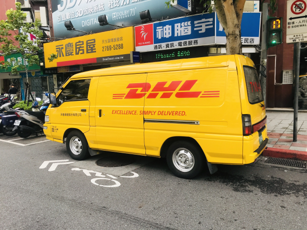 Gelber DHL-Lieferwagen auf der Straßenseite geparkt, umgeben von Gebäuden, Bäumen, Schildern, Masten, Verkehrssignalen und einer Person auf dem Gehweg.