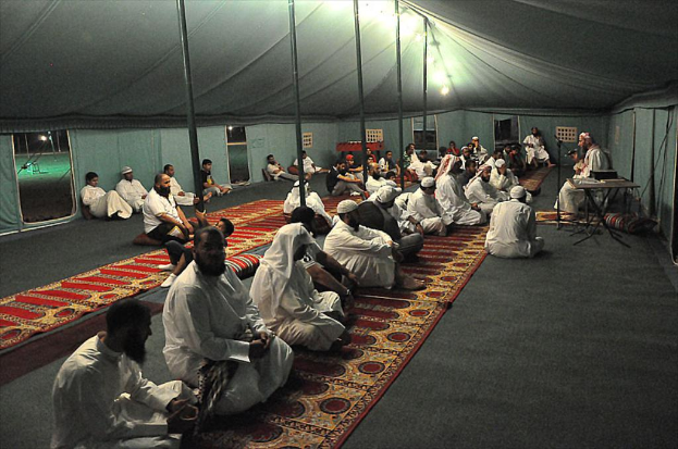 Eine Gruppe von Menschen, die auf dem Boden in einem Lager sitzen, mit einer Person, die rechts auf einem Stuhl sitzt, einem Mikrofon vor ihnen, einem Tisch neben ihnen und einer Lampe oben im Bild.