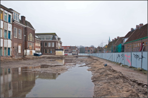 Eine überflutete Stadtstraße mit Wasser auf dem Boden, ein Zaun auf der rechten Seite, Gebäude mit Fenstern auf der linken Seite, Bäume im Hintergrund und ein Himmel darüber.