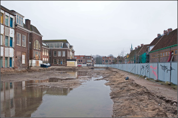 Eine überflutete Stadtstraße mit Wasser auf dem Boden, ein Zaun auf der rechten Seite, Gebäude mit Fenstern auf der linken Seite, Bäume im Hintergrund und ein Himmel darüber.
