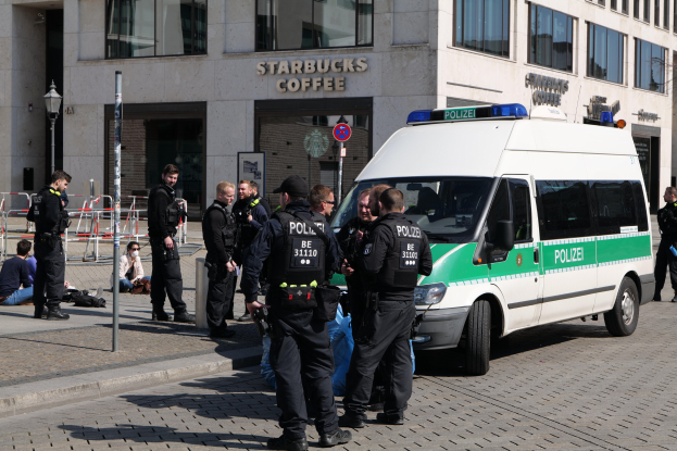 Eine Gruppe von Polizisten steht vor einem Starbucks-Café, rechts ein Van und links ein paar Menschen, vor einem Gebäude mit Fenstern, einer Tafel, einem Laternenpfahl und einem Zaun.