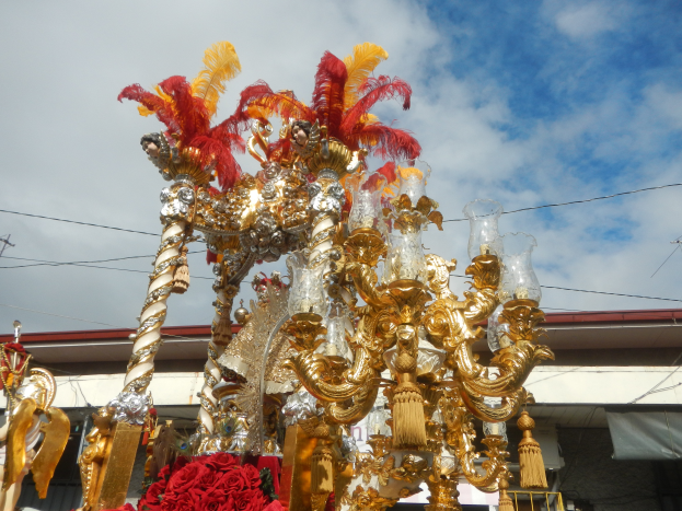 Ein großes goldenes und rotes Festwagen mit Blumen und anderen Dekorationen bei einem Karnevalsumzug, mit einem Gebäude, Strommasten mit Drähten und einem bewölkten Himmel im Hintergrund.