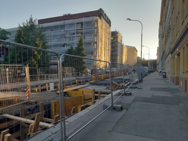 Stadtstraße mit einer Baustelle umgeben von Gebäuden, Straßenlaternen, Straßenlaternen, Fahrzeugen, Absperrpoller, Bäumen und einem klaren blauen Himmel.