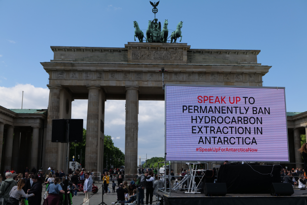 Eine Gruppe von Menschen steht vor dem Brandenburger Tor in Berlin, Deutschland, mit einer Frau auf einer Bühne rechts, Rednern und einem Bildschirm mit dem Text "Sprecht euch aus, um die permanente Verbrennung von Kohlenwasserstoffen in der Antarktis zu verbieten". Im Hintergrund ist das Brandenburger Tor mit seinen Säulen und Statuen sowie Bäume und ein bewölkter Himmel zu sehen.