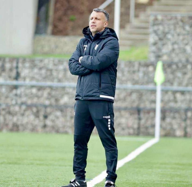Ein Mann in schwarzer Jacke und Hose steht auf einem Fußballfeld mit verschränkten Armen und sieht aus wie ein Trainer während eines Trainings, mit einem Zaun, einer Treppe und einer Wand im Hintergrund.