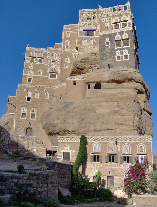 Ein Schloss mit Fenstern und T√ľren, umgeben von Pflanzen, unter einem blauen Himmel.