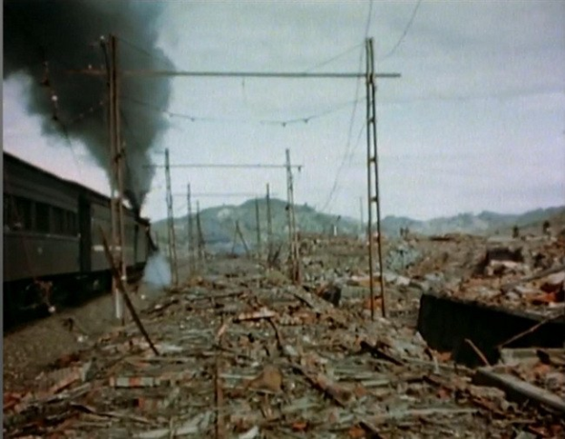 Ein Zug fährt durch ein zerstörtes Gebiet mit Rauch, der aus ihm aufsteigt, umgeben von Strommasten und -leitungen, mit Hügeln und einem klaren Himmel im Hintergrund und verstreuten Schrottteilen auf dem Boden.