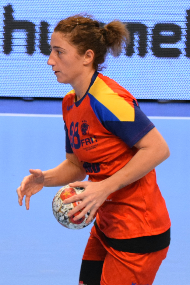 Eine Frau in orange-blauer Uniform hält einen Fußball, im Hintergrund ist ein Bildschirm mit Text zu sehen.