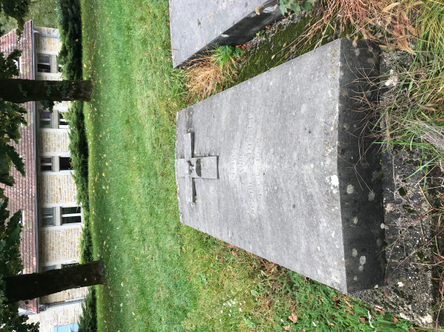 Ein Friedhof mit einem Grab in der Mitte, umgeben von Gras, Pflanzen und Bäumen, mit einem Gebäude mit Fenstern im Hintergrund.