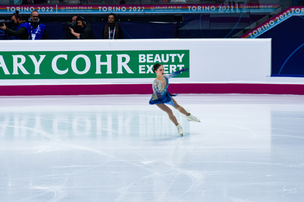 Eine Frau in einem blauen Kleid fährt auf einem Eisstadion Schlittschuh, während eine Menge mit Kameras und einem Schild mit der Aufschrift 'Tessa Virtue und Scott Moir' im Hintergrund steht.