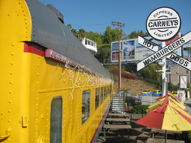 Ein mobiles Kantinenfahrzeug ist in der Nähe von Bänken und Treppen geparkt, mit Schirmen, Pflanzen und einem Geländer in der Nähe. Fahrzeuge, ein Pfahl, Bretter und Werbetafeln sind ebenfalls sichtbar, zusammen mit einem Gebäude, Bäumen und verschiedenen Gegenständen unter einem klaren Himmel.