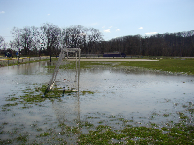Fußballtor in einem überfluteten Feld mit umgebender Wiese, Zaun, Bäumen, Häusern und einem bewölkten Himmel.