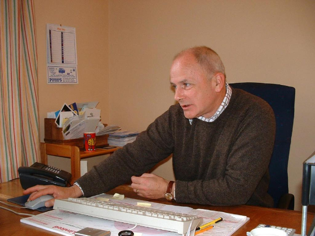 Ein Mann sitzt an einem Tisch mit einem Telefon, einer Tastatur, einem Buch, einem Stift und einem Taschenrechner, mit einer Wand, einem Kalender und einem Vorhang im Hintergrund und einer Tasse und Papieren auf dem Tisch.