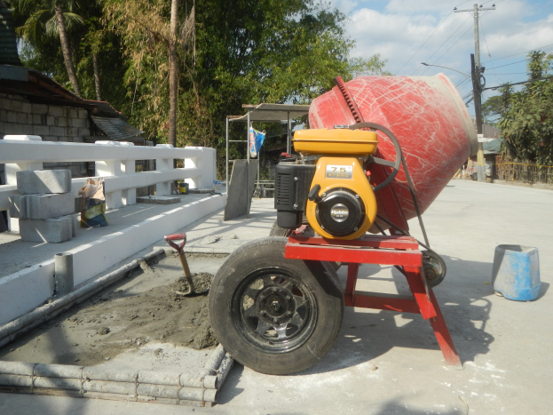 Betonmischer auf einem Zementlastwagen umgeben von Baustoffen, einem Haus, Bäumen, Strommasten und einem bewölkten Himmel.