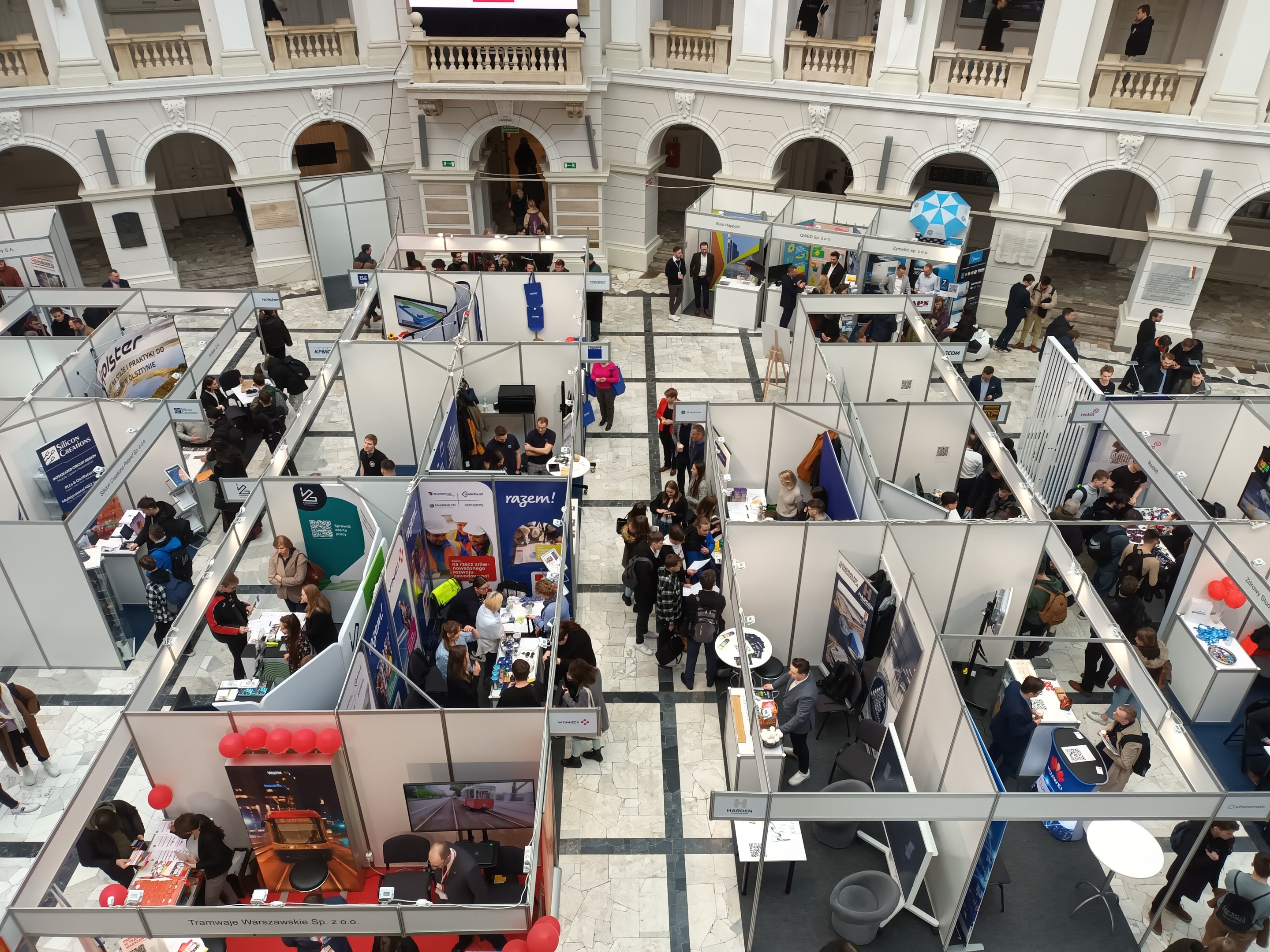 Eine große Gruppe von Menschen auf einer internationalen Fachmesse mit Ständen, Bannern, Tischen, Stühlen und Ballons in einem Raum mit architektonischen Bögen, Säulen, Geländern und einem Bildschirm im Hintergrund.