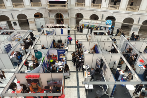 Eine große Gruppe von Menschen auf einer internationalen Fachmesse mit Ständen, Bannern, Tischen, Stühlen und Ballons in einem Raum mit architektonischen Bögen, Säulen, Geländern und einem Bildschirm im Hintergrund.