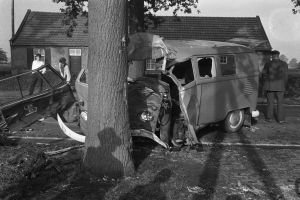 Eine schwarz-weiße Szene eines verunglückten Autos am Straßenrand, mit einem Baum im Vordergrund, einigen Menschen in der Nähe und Häusern, Bäumen und einem Mast im Hintergrund.