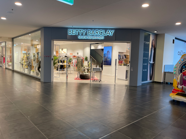 Das Innere eines Betty Barclay-Ladens in einem Einkaufszentrum mit Schaufensterpuppen in Kleidern, hängenden Kleidern, Tischen, Pflanzen und einem Spielzeugfahrzeug auf der rechten Seite, mit einer Tafel mit der Aufschrift "Betty Barclay Selected Brands" oben.