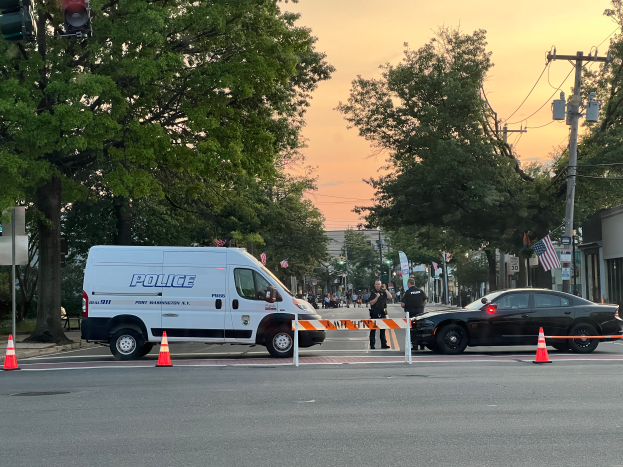 Ein Polizeiwagen steht an der Seite einer Straße neben einer Ampel, umgeben von Verkehrskegeln, Pfosten, Schildern und anderen Gegenständen, mit Gebäuden, Bäumen und Fahnen im Hintergrund, nach einem Schusswaffen Vorfall in Port Washington.