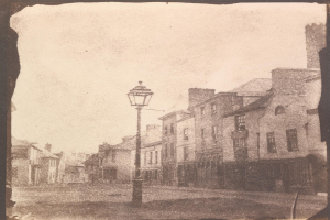 Altes Schwarz-Weiß-Foto einer Straßenlaterne an einer Kreuzung, flankiert von Gebäuden unter einem klaren Himmel.