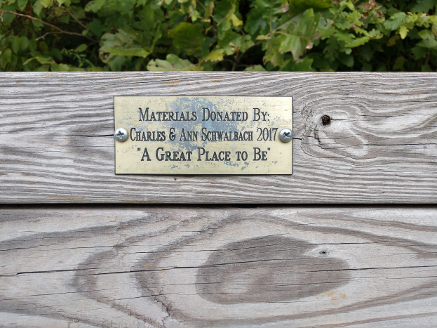 Holzbank mit einer Plakette, auf der "Materialien gespendet von Charles & Ann Schwabach 2017: Ein schöner Ort" steht, umgeben von üppigen grünen Pflanzen.