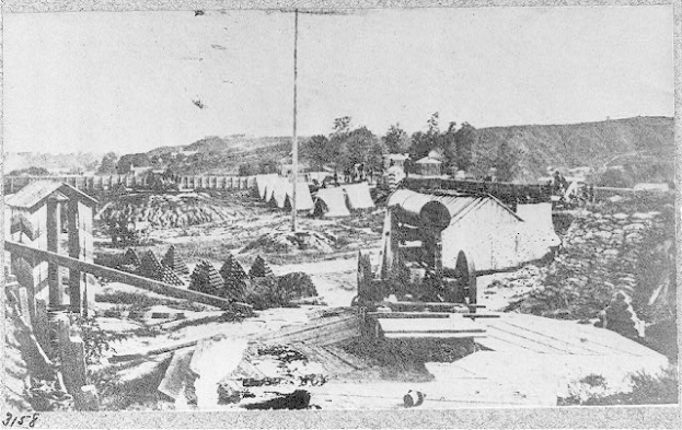 Schwarzes und weißes Foto einer Baustelle mit einer Maschine im Vordergrund, Schuppen, Bäumen, Hügeln und Himmel im Hintergrund, mit Text unten, der "Die Bauarbeiten an einer neuen Brücke in der Stadt San Francisco, Kalifornien" lautet.