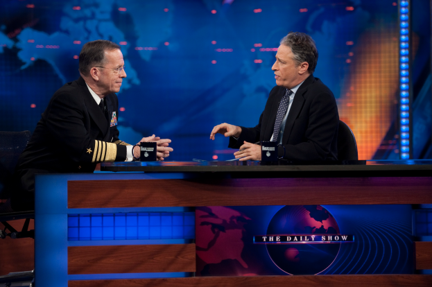 Zwei Männer in schwarzen Blazern, Hemden und Krawatten sitzen an einem Tisch mit Mikrofonen dazwischen vor einem TV-Bildschirm und einem blauen Hintergrund.