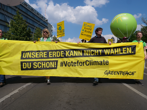 Eine Gruppe von Menschen steht auf einer Straße und hält ein gelbes "Stimme für das Klima"-Plakat, mit Bäumen, einem Gebäude, einem Pfahl, einem Turm und einem klaren blauen Himmel im Hintergrund und einem grünen Ballon auf der rechten Seite.