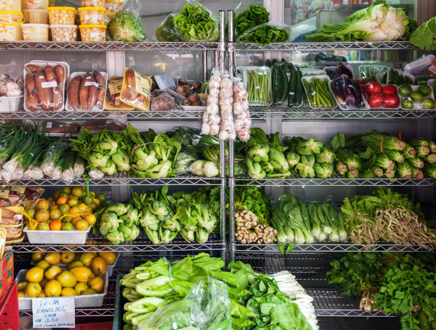 Ein Gang in einem Supermarkt mit frischem Gemüse und Obst, verpackten Artikeln in Plastikfolien, Kartons, einer Texttafel und einem Glasfenster im Hintergrund.
