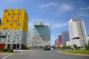 Eine Stadtstraße mit hohen Gebäuden im Hintergrund, Straßenlaternen, Autos, Informationsschildern, Büschen, Bäumen und einem bewölkten Himmel, die einzigartig gestalteten Gebäude der Universität Rotterdam.