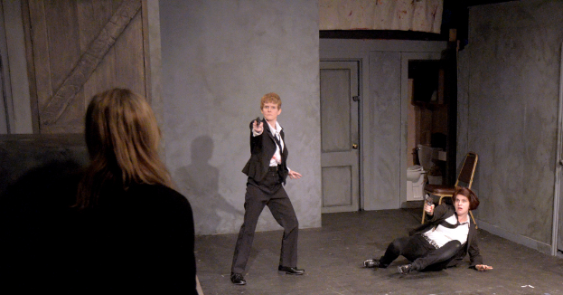 Zwei Frauen in schwarzen Anzügen, eine sitzt auf dem Boden und hält eine Waffe, die andere steht und hält eine Waffe, mit einer dritten Frau, die daneben steht, in einem Raum mit einem Stuhl, einer Toilette und einer hölzernen Tür.