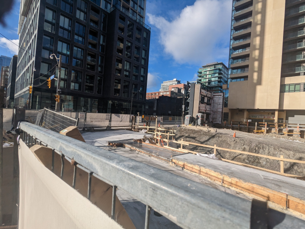 Baustelle in Toronto, Ontario mit einem großen Bauwerk im Bau, umgeben von Absperrungen, Pfählen, Verkehrssignalen und Schildern, unter einem bewölkten Himmel.