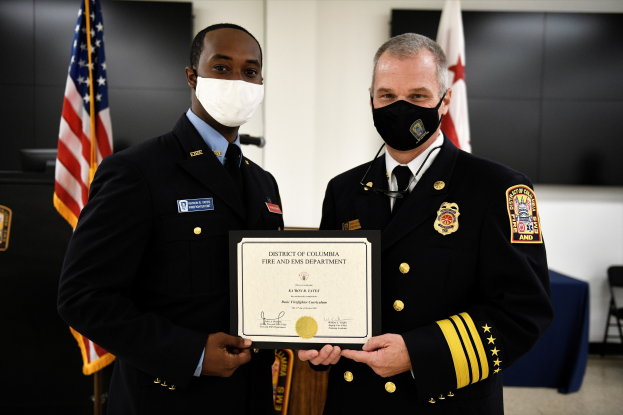 Zwei Männer in Feuerwache-Uniformen stehen nebeneinander, tragen Masken und halten ein Zertifikat, mit Flaggen und Möbeln im Hintergrund; der Mann links ist der Feuerwehrchef des Bezirks Washington, der einen Preis erhält.