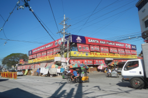 Lebendige Straßenszene mit einem Lastwagen, mehreren Fahrzeugen, Fußgängern, einem Strommast mit Drähten, Bäumen und einem klaren blauen Himmel.