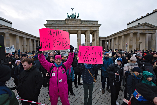Eine Gruppe von Menschen steht vor dem Reichstagsgebäude in Berlin und hält Schilder, die gegen die Regierungskampagne gegen Neonazis protestieren, mit Gebäuden und Statuen im Hintergrund unter einem klaren blauen Himmel.
