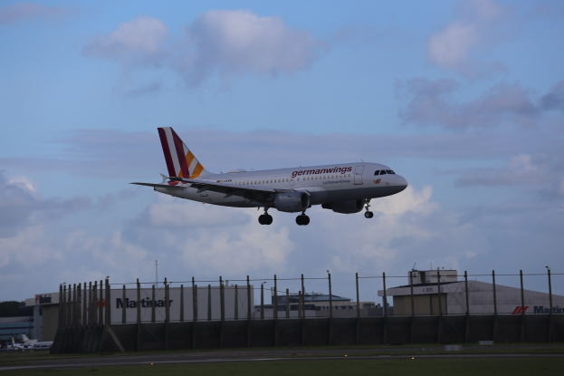 Germanwings Airbus A320-200 beim Start von Frankfurt Airport, mit Gras, einem Zaun, Gebäuden, Bäumen und einem bewölkten Himmel im Hintergrund.