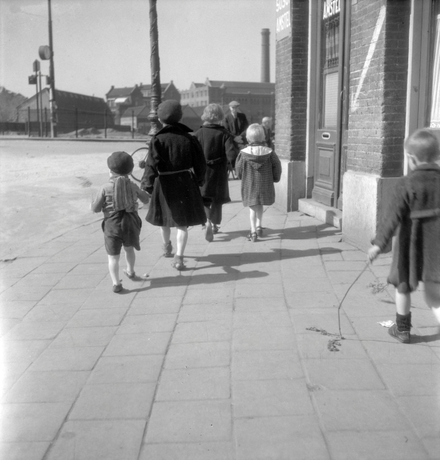 Eine Gruppe von Kindern, die auf einem Gehweg gehen, mit einem Gebäude auf der rechten Seite, das eine Tür und Fenster hat, und Masten, Gebäuden, Bäumen und einem klaren Himmel im Hintergrund.