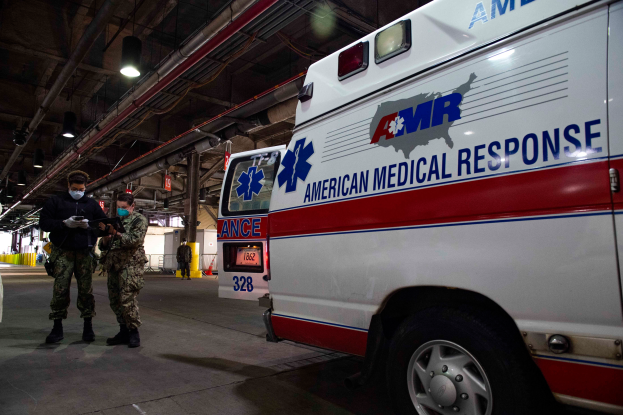 Ein Rettungswagen der American Medical Response (AMR) steht in einem Gebäude, vor ihm stehen zwei maskierte Personen, links ein Fahrzeug und im Hintergrund mehrere Personen, Lichter, Rohre und andere Gegenstände.