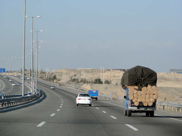 Ein Holzlaster mit einer großen Ladung Holz fährt auf einer Autobahn mit Schutzplanken, Laternen, Schildern, Bäumen und Sand, mit Hügeln und einem klaren blauen Himmel im Hintergrund.