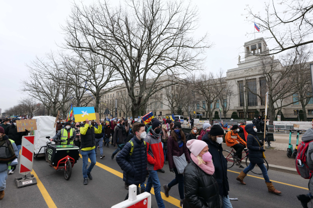 Eine große Gruppe von Menschen marschiert auf einer Straße in Washington, D.C. und hält Schilder und fährt Fahrräder, mit Gebäuden, Bäumen und einem klaren blauen Himmel im Hintergrund.